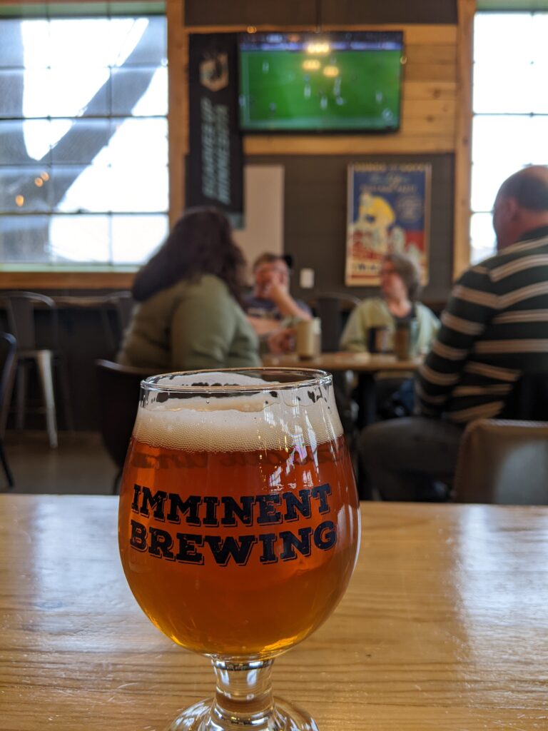 A beer labeled "Imminent Brewing" on a table, with people seated at a table and a TV showing a soccer game in the background.